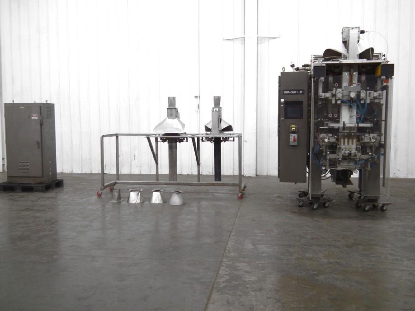 A packaging machine setup in a large, empty industrial room with white panel walls showcases surplus industrial food and beverage equipment, with various machine parts and metal funnels arranged on the floor and metal frame.