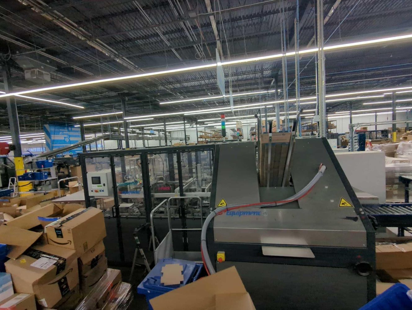 A busy warehouse or factory floor with conveyor belts, machinery, and stacks of cardboard boxes showcases an industrial packaging operation featuring Packable LLC - Pristine E-Commerce Fulfillment Equipment Auction: Sealed Air I-Pack Void Reduction System.
