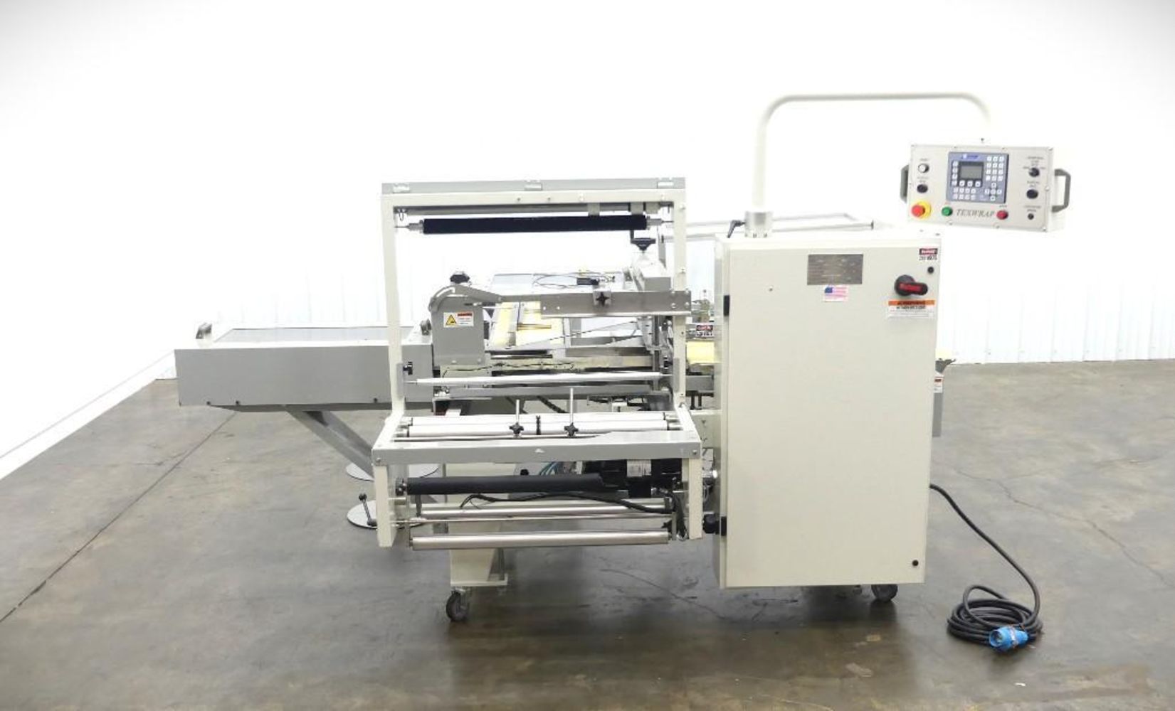 A white industrial packaging machine with conveyor belts, control panel, and exposed mechanical parts sits on a gray concrete floor in a plain room—featured at the Monthly SIGMA Showroom Floor Multi-Industry Auction. An electrical cord lies coiled beside it.
