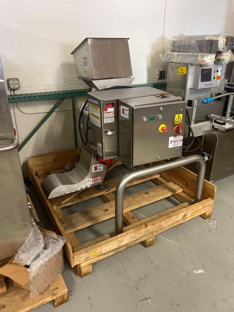 A stainless steel industrial food processing machine sits on a wooden pallet in a warehouse—part of the Pilot Plant Liquidation: Food Processing and Packaging Equipment, featuring pristine assets of a Fortune 100 company. Control buttons and safety labels are visible.