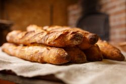 Four freshly baked baguettes stacked on a rustic cloth, with a brick oven blurred in the background—capturing the warmth and craftsmanship you’ll find at our Quarterly Bakery and Snack Food Processing and Packaging Equipment Auction.