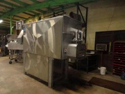 A large stainless steel industrial machine with a control panel stands in a dimly lit factory, ready for the Monthly Sigma Showroom Floor Multi-Industry Auction. Metal racks, a bucket, and other equipment fill the background.