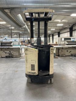 A used Crown forklift with visible scratches and worn paint is parked in a large industrial warehouse, surrounded by machinery and equipment from The Bindery - Industrial Bindery Equipment Auction.