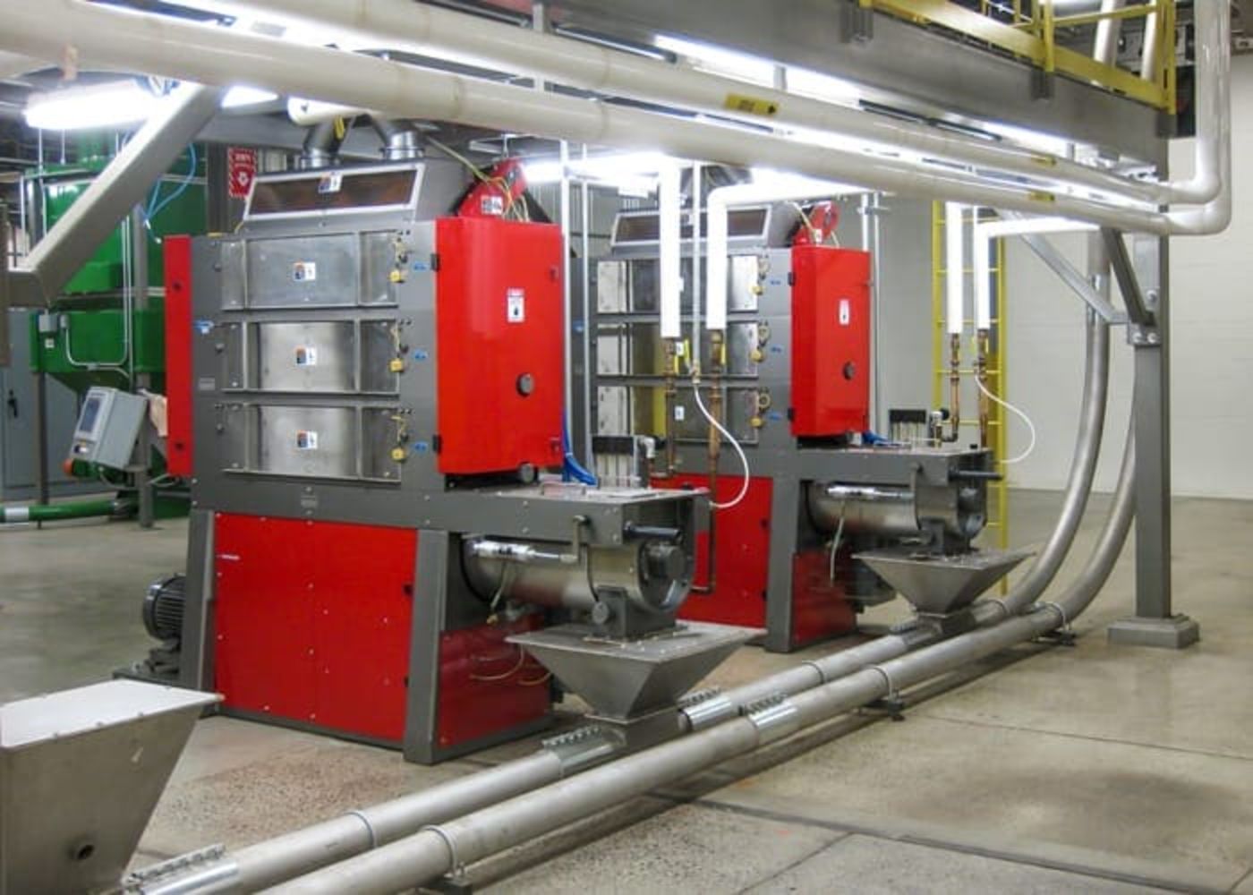 Two large industrial machines with red and silver casings, surplus processing and packaging equipment from a Fortune 500 snack food manufacturer, are positioned side by side in a factory setting surrounded by metal pipes and concrete flooring.