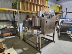 A large industrial stainless steel machine stands on a metal frame in a warehouse, surrounded by shelves with boxes and other equipment. The floor is concrete and the lighting is bright—ideal for the auction for ongoing operation of a popcorn and fudge producer.
