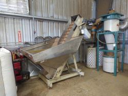A metal industrial machine with a slanted conveyor and hopper stands in a workshop with corrugated metal walls, featured in the Hemp Depot: Hemp Processing Equipment Auction, alongside dust collection equipment nearby.
