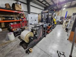 A large industrial webbing printing machine sits in a spacious warehouse, surrounded by shelves of boxes and equipment, ready for processing at the Drilling Technical Services: Webbing Printing Equipment Auction.