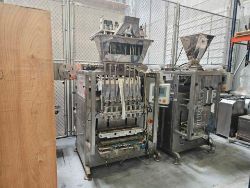 Two large industrial packaging machines—ideal for powder and liquid stick packs—stand side by side in a warehouse with concrete floors and metal fencing, showcased at the Nutraceutical Equipment Auction.