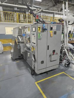 Industrial machine with control panels and cables in a factory setting; yellow railings and equipment, including Piston Filler Cappers, are visible in the background, with the floor marked by yellow lines.