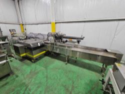 A stainless steel industrial machine with conveyor belt stands in a factory room with green flooring and white paneled walls, offered as part of Bektrom Foods: Excess Equipment Sale for the Food and Beverage Industry. Ideal for food processing or packaging.