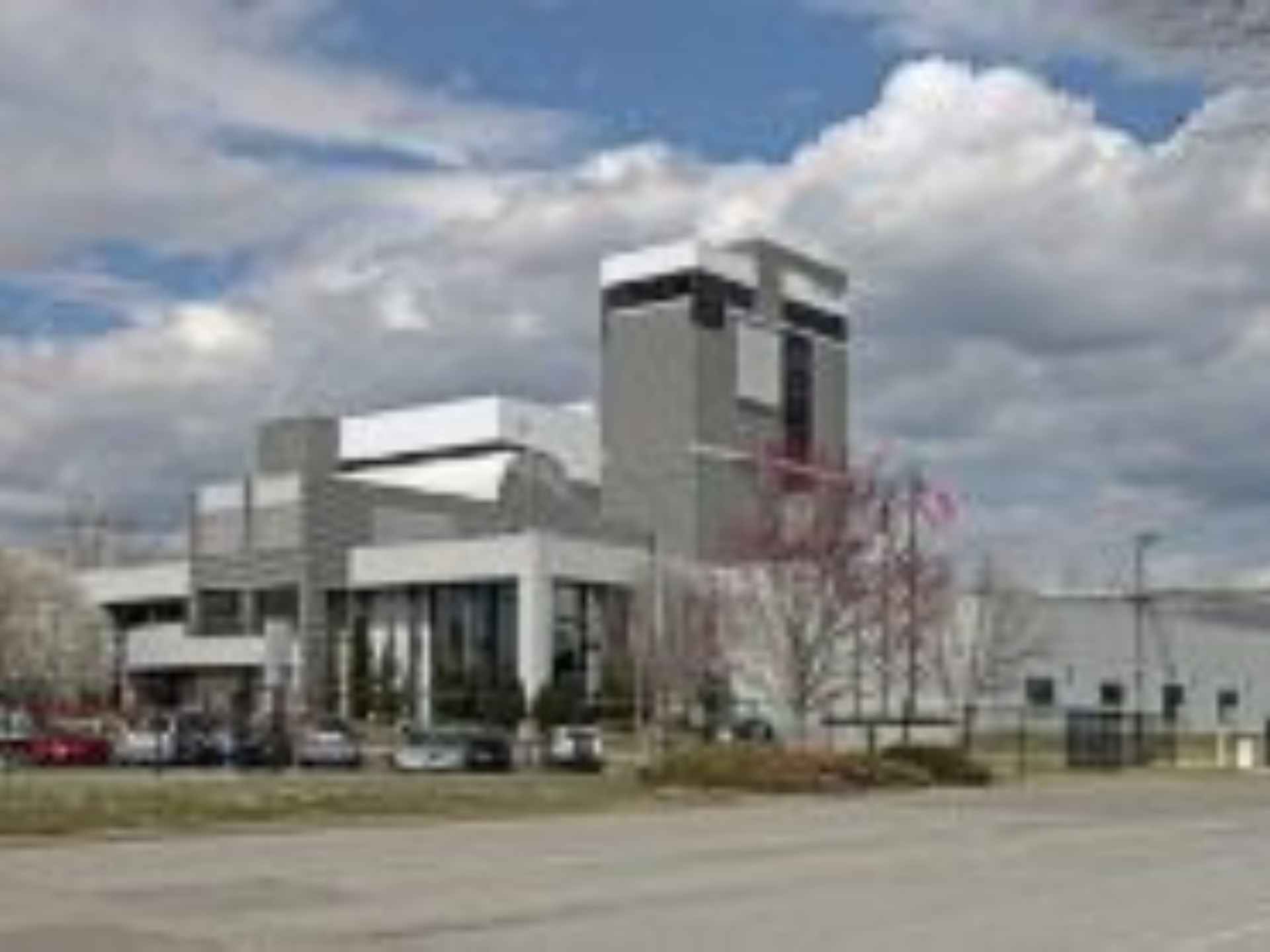 A large, modern industrial building with tall sections and angular architecture, surrounded by parked cars and a few leafless trees under a partly cloudy sky—site of industrial and support equipment from a major coffee production facility auction.