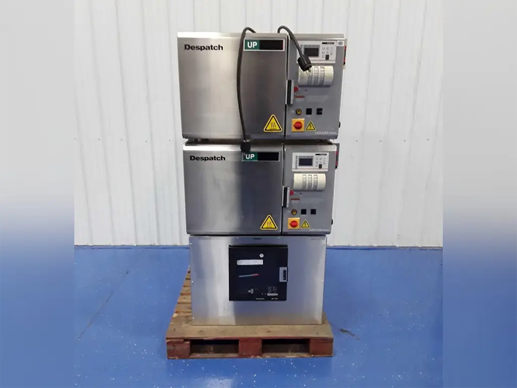 Two stacked industrial Despatch ovens with control panels and warning symbols, set on a wooden pallet on a blue floor against a white wall—ideal for those seeking quality at a Surplus Lab Equipment Auction.