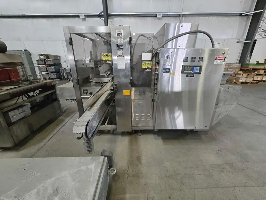 A large industrial packaging machine with a conveyor belt stands in a warehouse with concrete floors and metal shelving, surrounded by various equipment and boxes.