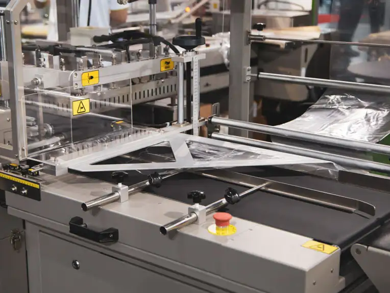 Automatic packaging machine wrapping an item with clear plastic film, featuring safety warning labels, metal bars, and control buttons—ideal for PPT Research: Plant Support Equipment and Laboratory Equipment Sale applications.